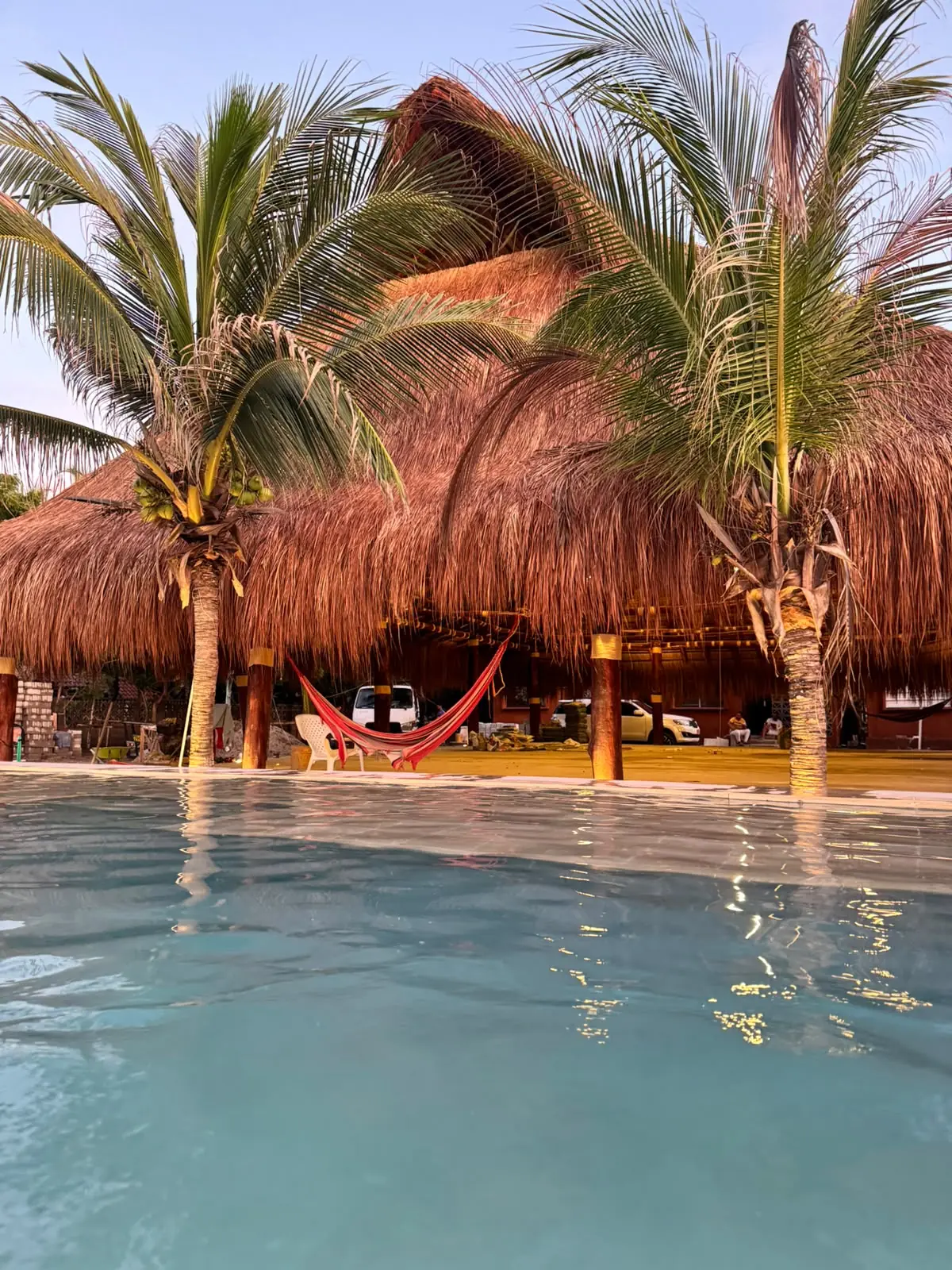 Gran palapa de palma con cocoteros y una hamaca roja junto a la piscina de Mokano